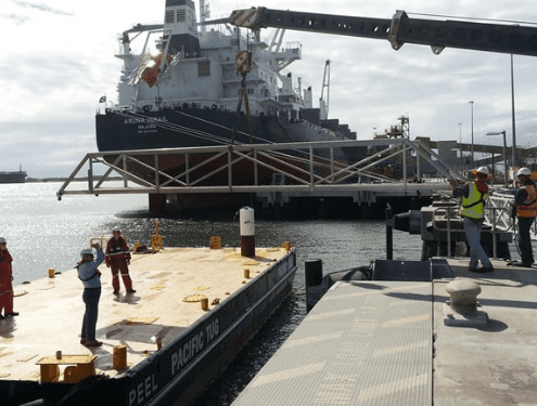 workers at a dock maneuvering a large metal structure with a crane near a boat